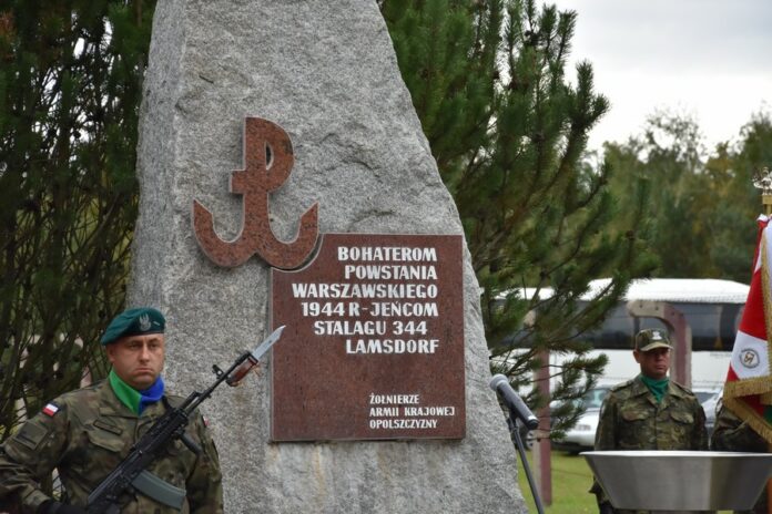 Pomnik poświęcony obecności powstańców warszawskich w Lamsdorf. Kamień zwieńczony krzyżem. Tablica pamiątkowa. Ptzy pomniku stoją żołnierze.