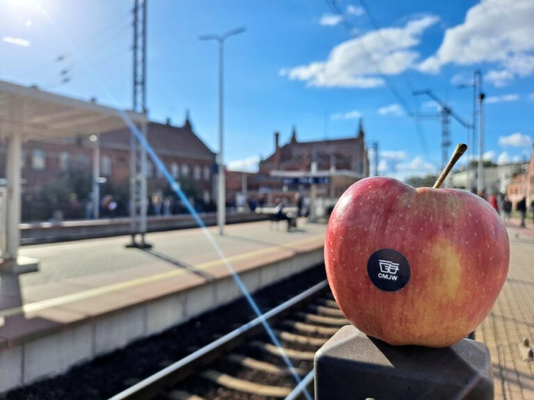 Jabłko sfotografowane na peronie. Widać tory itrakcje kolejową oraz budynki dworca. Na jabłku jest mała czarna naklejka z literam CMJW